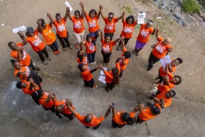 Participants in orange shirts form a circle outdoors and raise their hands.