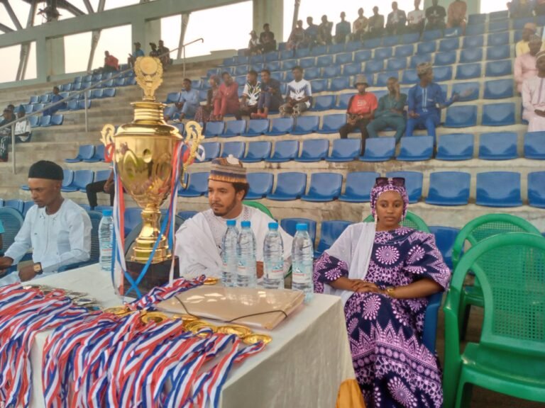 A trophy stands on a table in front of seated event organizers.